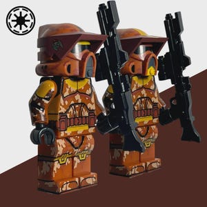 May include: Two orange and brown Lego figures in camouflage armor, helmets, and black rifles. The figures have yellow accents and are standing side-by-side. A white and black symbol is in the upper left corner.