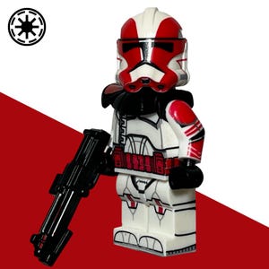 May include: A white and red plastic toy soldier minifigure featuring a detailed helmet with a red visor, a black neck collar, and red arm markings. The figure holds a black plastic blaster rifle and displays a red belt and leg armour details.