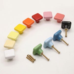 May include: A selection of square ceramic cabinet knobs in a range of colours, including white, yellow, orange, red, and blue. Each knob has a brass screw for fitting. The knobs are arranged in a semi-circle.