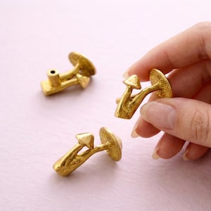 Könnte beinhalten: Goldfarbene Pilz-Schrankknöpfe. Die Knöpfe haben ein detailliertes Design mit einem abgerundeten Hut und einem Stiel. Die Knöpfe sind aus Metall und haben eine glänzende Oberfläche. Die Knöpfe sind für den Einsatz an Schränken oder Schubladen konzipiert.