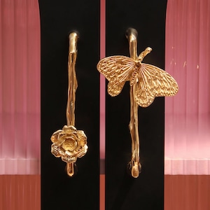 May include: Ornate gold door handles. One is shaped like a flower, the other a butterfly. They are mounted on black panels, with a pink, textured backdrop. These handles offer a luxurious, nature-themed aesthetic.