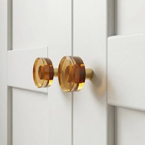 May include: Two amber-colored cabinet knobs with a circular design and brass hardware. The knobs are mounted on a white cabinet door. The knobs have a transparent, layered appearance, adding a touch of elegance to the furniture.