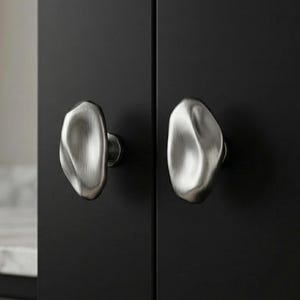 Puede incluir: Dos tiradores de armario de níquel cepillado con un diseño orgánico en forma de guijarro. Los tiradores están montados en una puerta de armario negra, ofreciendo una estética moderna y minimalista. El herraje tiene una superficie lisa y texturizada.