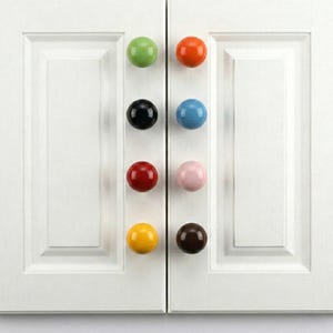 Könnte beinhalten: Eine Sammlung von acht bunten Keramik-Schrankknöpfen in verschiedenen Farbtönen, darunter Grün, Orange, Schwarz, Blau, Rot, Rosa, Gelb und Braun. Die Knöpfe sind rund und an weißen Schranktüren befestigt.
