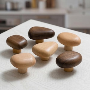 Puede incluir: Seis pomos de madera para armarios en varios tonos de marrón y madera natural. Los pomos tienen forma redondeada, similar a guijarros, y están montados sobre bases cortas y cilíndricas. Los pomos están dispuestos sobre una superficie blanca.