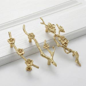 May include: Four gold-colored cabinet pulls in a floral branch design. Each pull features a branch-like structure with delicate flower details. The pulls are mounted on a white cabinet door, showcasing their decorative nature.