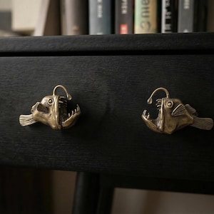 May include: Two brass-colored, anglerfish-shaped drawer pulls are mounted on a black wooden surface. The pulls have open mouths with visible teeth, a fin, and a light fixture above the head. The drawer pulls are decorative and add a unique touch to furniture.