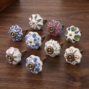 May include: A collection of nine ceramic cabinet knobs with floral designs. The knobs are white with blue, red, and green patterns, and have brass-colored hardware. The knobs are arranged on a dark wooden surface.