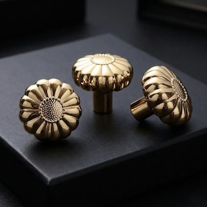 May include: Three gold-colored cabinet knobs with a sunflower design. The knobs have a fluted edge and a textured center, giving them a vintage look. The knobs are made of metal and are likely for furniture.