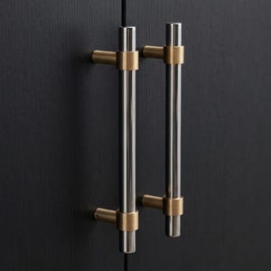 May include: Two cabinet pulls with a modern design. Each pull features a brushed nickel bar with brass-colored accents. The pulls are mounted on a dark wood cabinet door.