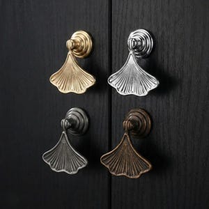 May include: Four cabinet pulls in gold, silver, dark gray, and bronze. Each pull features a fan-shaped design with a circular base, mounted on a dark wood surface. The pulls are arranged in a square, showcasing their different metallic finishes.
