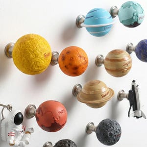 May include: A collection of decorative cabinet knobs and pulls, each designed to resemble a planet or celestial object. The knobs are in various colors, including yellow, orange, blue, and brown, with labels such as "Sun", "Mars", and "Jupiter". A small astronaut figure is also included.