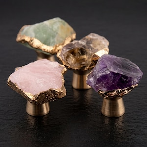 May include: Four decorative cabinet knobs with natural stone faces in shades of green, brown, pink, and purple. Each stone is edged with gold and mounted on a brass base. The knobs are arranged on a dark surface.