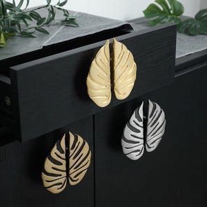 Op de afbeelding: Close-up van drie kastgrepen in de vorm van gespleten Monstera-bladeren. Twee zijn goudkleurig en één is zilverkleurig. De grepen zijn gemonteerd op een zwarte kast met een open lade. Decoratieve hardware.