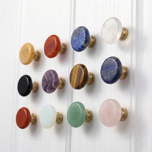 May include: Twelve cabinet knobs with brass fittings. The knobs are made from polished gemstones in various colours, including yellow, red, blue, clear, black, purple, brown, white, and pink. They are mounted on a white surface.