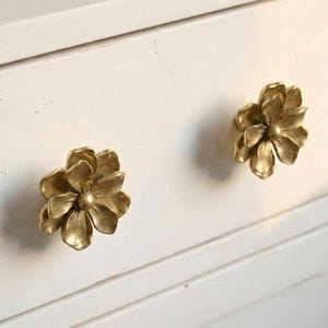 May include: Two brass-colored flower-shaped drawer pulls are mounted on a white cabinet. The drawer pulls are detailed with multiple petals and a central bud. The cabinet has a simple, clean design.