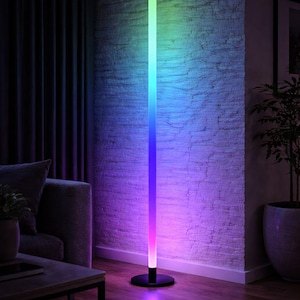 Puede incluir: Una lámpara de pie moderna con un degradado de colores, que va del verde al azul y al morado. La lámpara se apoya sobre una base circular negra y está colocada contra una pared de ladrillo blanco. La habitación tiene un ambiente oscuro.