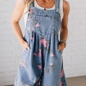 May include: Light blue denim overall shorts with a pink floral pattern. The shorts have a bib front with button closures, a front pocket, and side pockets. The rolled-up hem adds a casual touch. A white top is worn underneath.