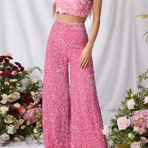 May include: A two-piece pink outfit featuring a sequined wide-leg pant and a strapless crop top with a rose-textured design. The outfit is a vibrant pink color, perfect for a special occasion.