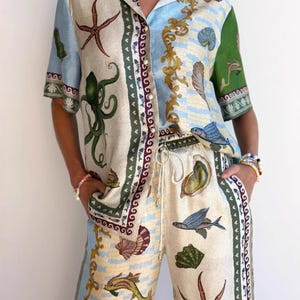 Puede incluir: Conjunto de dos piezas color crema, compuesto por una camisa abotonada y un pantalón con cordón. El estampado de temática marina incluye estrellas de mar, conchas y peces. La camisa tiene mangas cortas y cuello. El pantalón tiene un corte holgado.