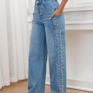 May include: Light blue denim jeans with a wide leg design. The jeans feature a series of small, white faux pearls along the side of the right leg. The jeans have a classic five-pocket design and a button and zipper closure.