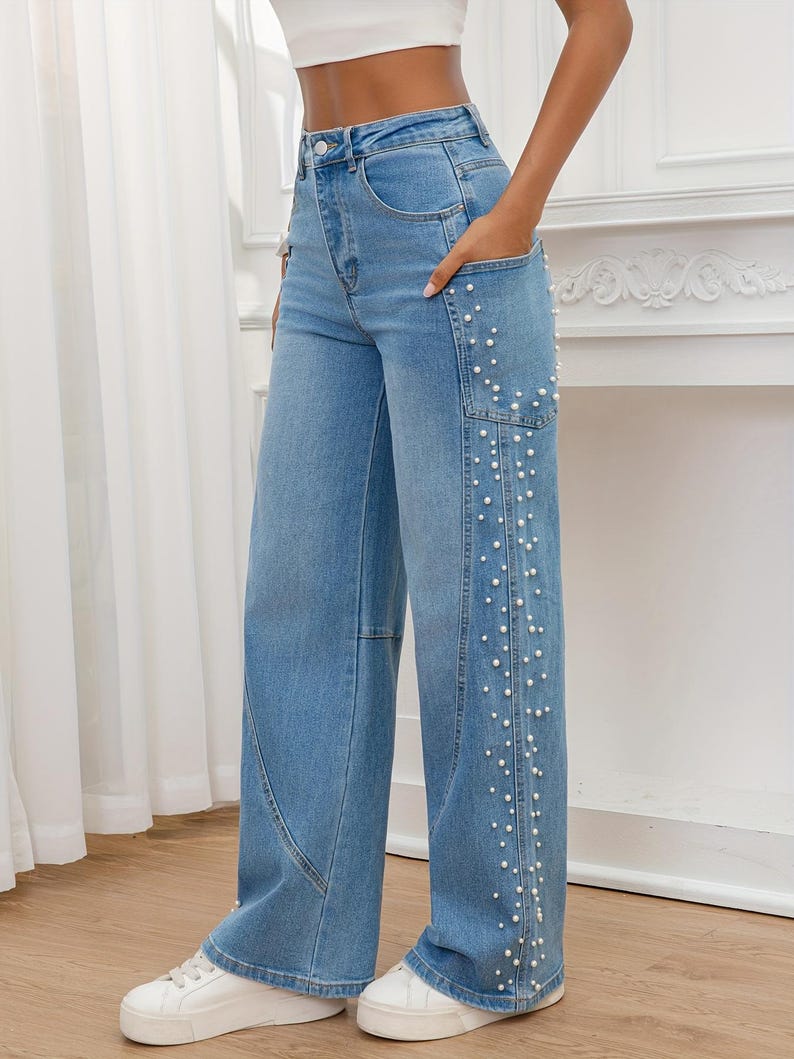 May include: Light blue denim jeans with a wide leg design. The jeans feature a series of small, white faux pearls along the side of the right leg. The jeans have a classic five-pocket design and a button and zipper closure.