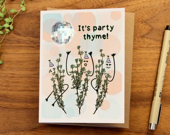 Puede incluir: Una tarjeta de felicitación con la frase "It's party thyme!". Presenta tres ramitas de tomillo antropomórficas con gorros de fiesta y brazos levantados. Una bola de discoteca y círculos pastel decoran el fondo. La tarjeta está sobre un sobre marrón.