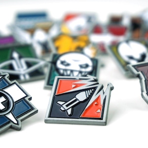 May include: A collection of colorful enamel pins with various designs. The pins feature a variety of shapes and colors, including red, black, white, and blue. Some pins have a military or tactical theme, with images of bombs, skulls, and crosses.