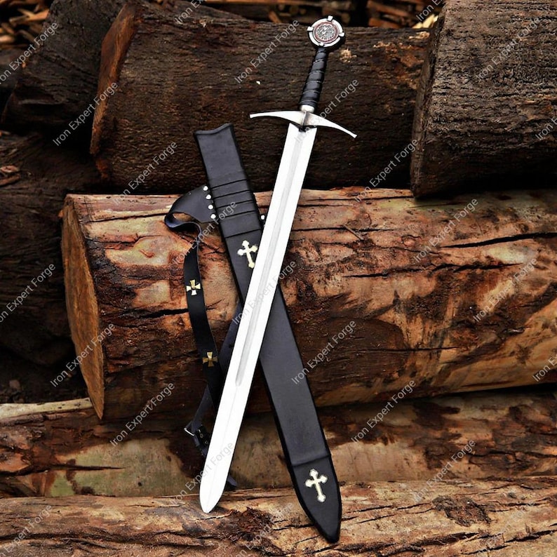 May include: A medieval sword with a silver blade and a black handle, resting in a black sheath. The sheath and sword handle feature gold cross designs. The sword is displayed against a backdrop of wooden logs.
