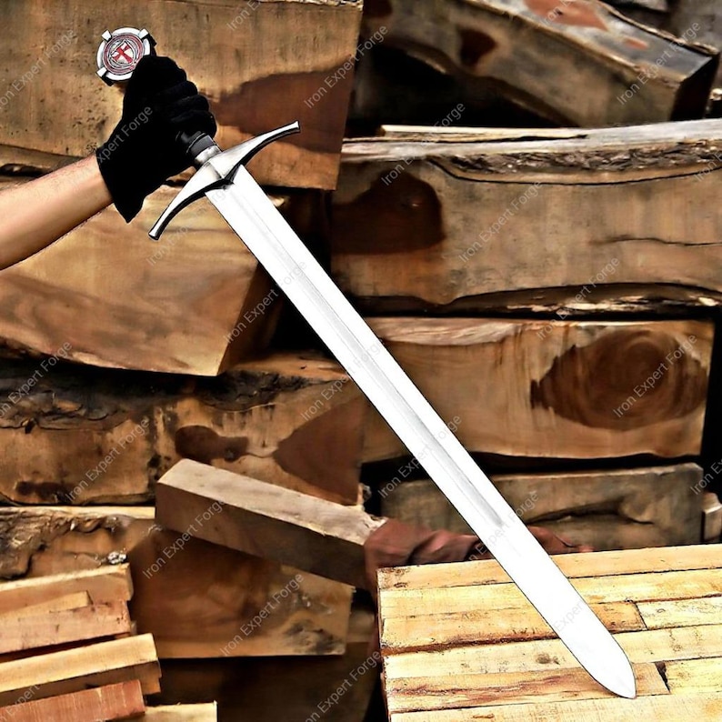 May include: A long, silver sword with a cross-shaped hilt and a decorative pommel. The blade is straight and sharp, reflecting the light. The sword is held against a backdrop of stacked wooden blocks, suggesting a medieval or fantasy theme.