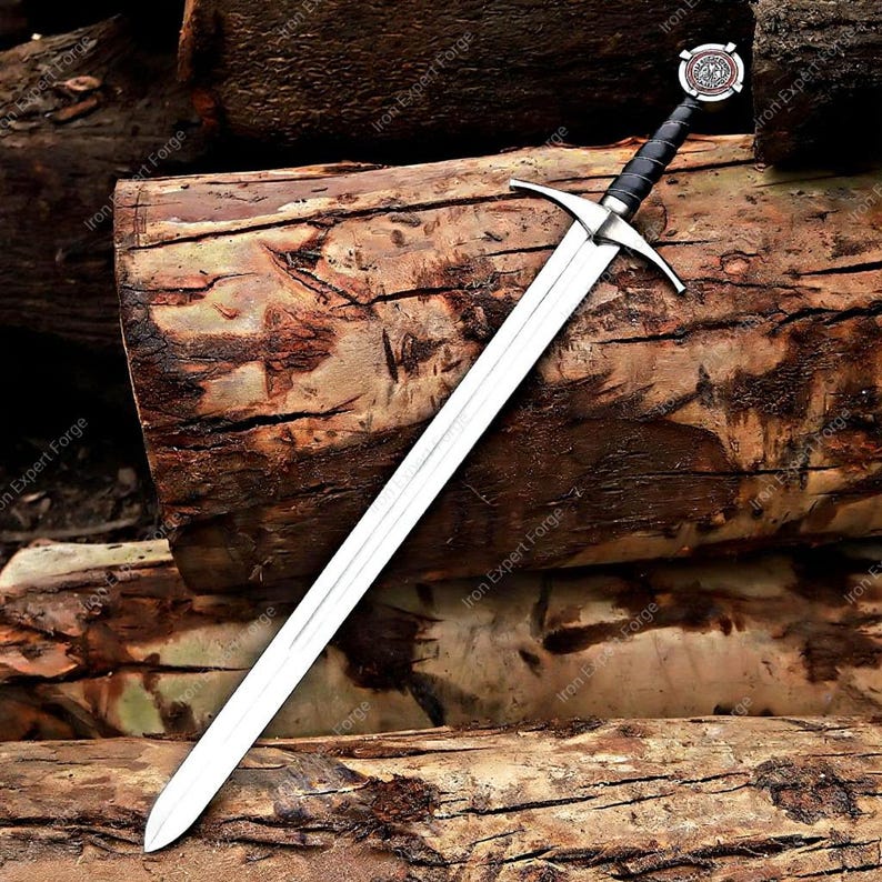 May include: A long, silver sword with a cross-shaped hilt and a black handle. The sword's blade is sharp and reflective, and the handle has a decorative emblem. The sword rests on a pile of wooden logs.