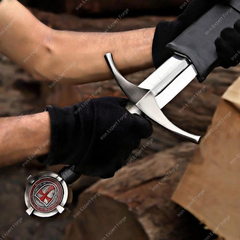 May include: A medieval sword with a silver blade and a black handle. The hilt features a decorative emblem with a red cross on a white shield. The sword is held by gloved hands, suggesting a focus on craftsmanship and historical design.