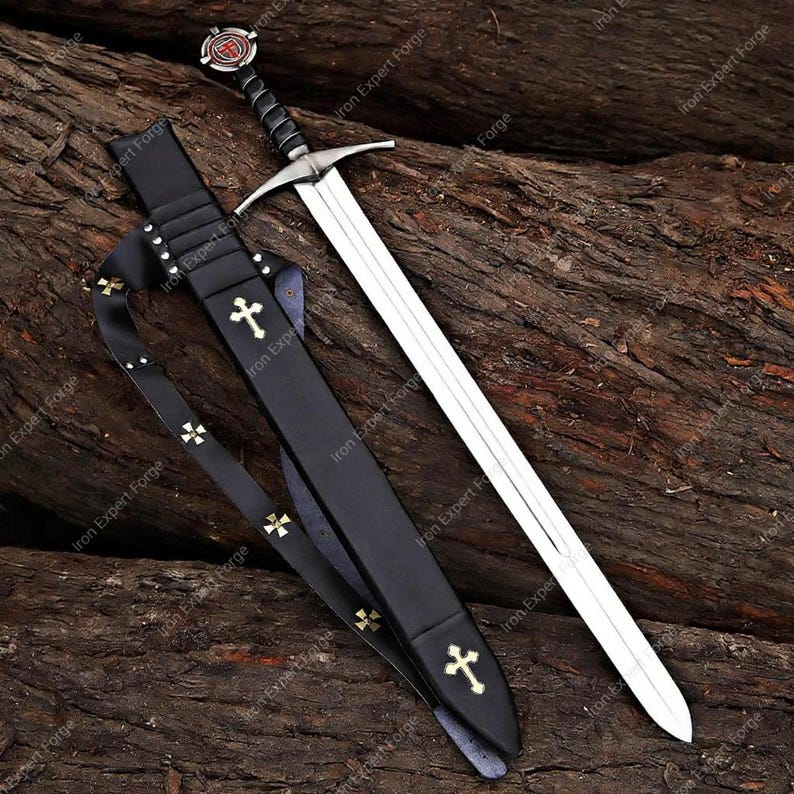 May include: A medieval sword with a silver blade and a black handle, resting on a wooden surface. The sword is accompanied by a black leather sheath adorned with gold crosses. The handle features a red and black emblem.