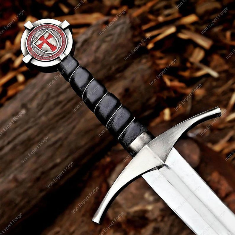 May include: A medieval-style sword with a silver blade and a black leather-wrapped handle. The pommel features a red and white emblem. The sword is displayed against a natural wood background.