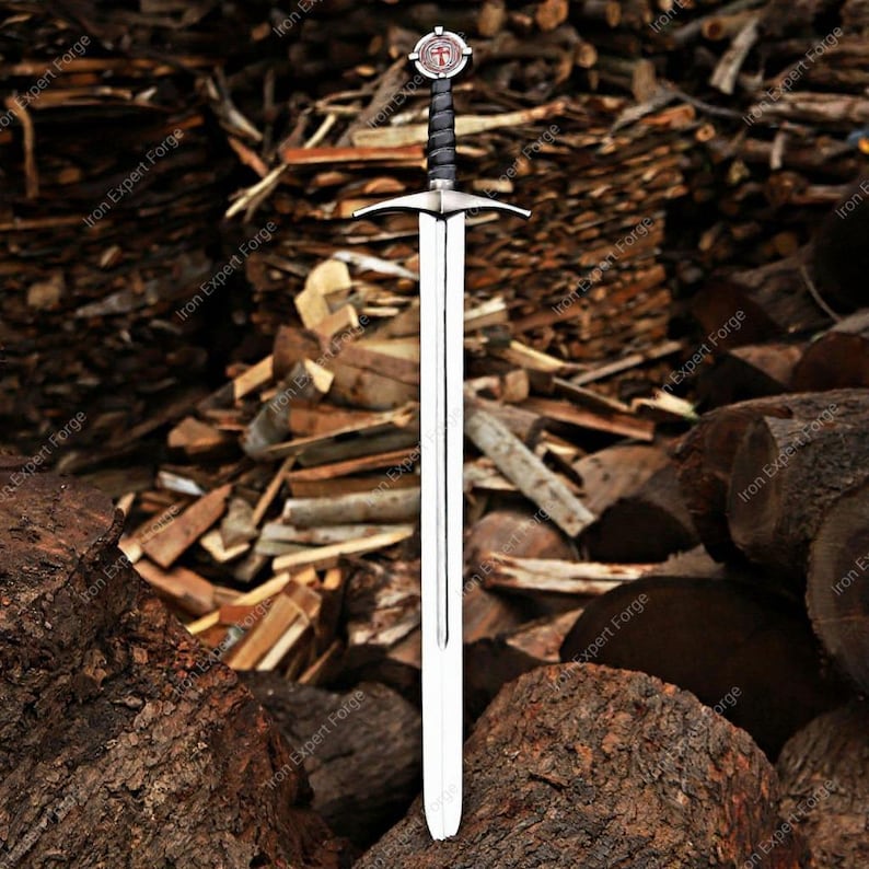 May include: A long, silver sword with a cross-shaped hilt and a black wrapped handle. The sword has a double-edged blade and a decorative emblem on the pommel. The background features a pile of wood logs.