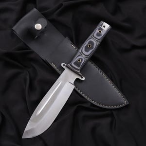 May include: A stainless steel hunting knife with a black and gray wooden handle and a black leather sheath. The knife has a wide blade and a full tang construction. The sheath has a snap closure.
