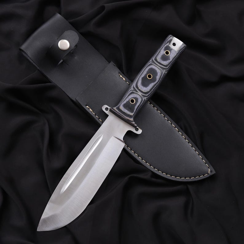 May include: A stainless steel hunting knife with a black and gray wooden handle and a black leather sheath. The knife has a wide blade and a full tang construction. The sheath has a snap closure.