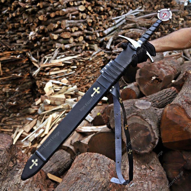 May include: A long, black sword with a silver crossguard and a black handle. The sword is in a black sheath with gold crosses. The sheath has a black strap with gold crosses. The sword's handle has a red and white emblem.