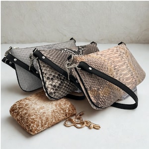 May include: Three small, textured handbags in shades of gray, brown, and tan, with black straps and silver hardware. A rectangular, textured pouch and a gold-colored metal accessory are in the foreground. The bags have a snakeskin pattern.