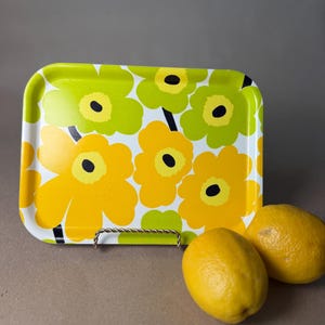 May include: Rectangular metal tray with a floral pattern in yellow, green, and black on a white background. Two yellow lemons sit beside the tray. The tray has a twisted metal handle.