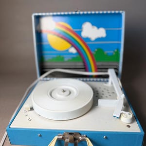 May include: Vintage blue and white portable record player with a cream-colored handle. The inside lid features a colorful rainbow and sun design. The turntable is white, and the player has a metal clasp.