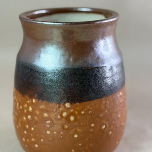 May include: A ceramic vase with a rounded body and a flared rim. The upper portion features a smooth, glossy brown glaze, while the middle section has a dark, textured black band. The lower half is a matte, light brown with a raised, bubbly texture.