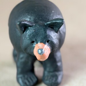 May include: A small, dark charcoal-colored bear figurine with a textured, matte finish. The bear has a rounded body, pointed ears, and a distinct, light tan-colored snout with a dark nose. The figure stands on four short, sturdy legs.