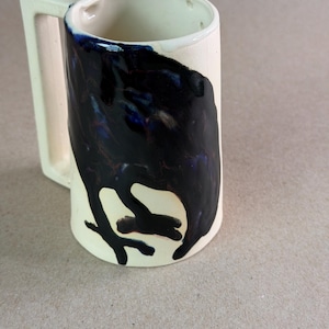 May include: A cylindrical ceramic mug with a cream-colored base and a glossy, dark navy blue glaze dripping down the front. The mug features a distinct, angular, rectangular handle extending from the side, providing a contrast to the rounded body.