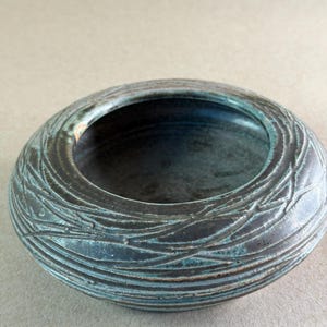 May include: A round, low-profile ceramic bowl featuring a dark, matte charcoal exterior with a textured, etched pattern of intersecting lines. The interior is glazed in a deep, mottled teal and blue, creating a contrast with the dark outer surface.
