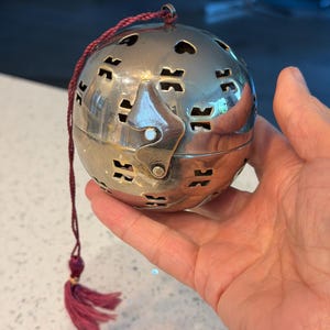 May include: A silver-colored, spherical container with decorative cut-out shapes. It has a hinged opening and a small, dark red tassel attached by a cord. The container is held in a hand.