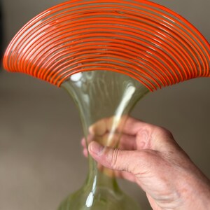 May include: A tall, green glass vase with a flared top, accented by a series of orange, curved lines. The vase has a bulbous base and a slender neck. The vase is held by a person's hand.