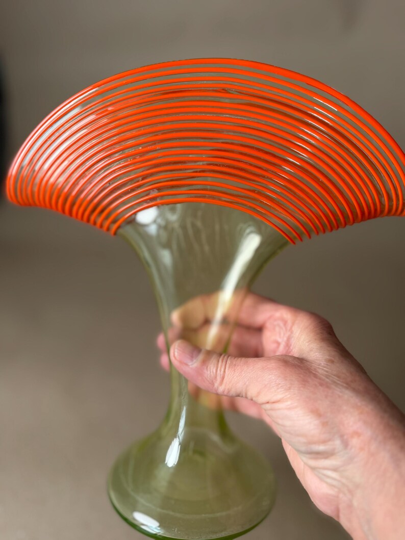 May include: A tall, green glass vase with a flared top, accented by a series of orange, curved lines. The vase has a bulbous base and a slender neck. The vase is held by a person's hand.