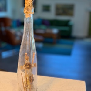 May include: A clear glass bottle with a cork stopper wrapped in natural fiber. Inside the bottle is a small, handcrafted figure made of natural materials. The bottle sits on a light-colored countertop.