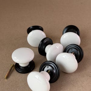 May include: A collection of six round cabinet knobs featuring smooth white ceramic tops and matte black metal bases. Each knob has a circular mounting plate and a central threaded hole. One separate gold-toned metal screw is placed nearby.